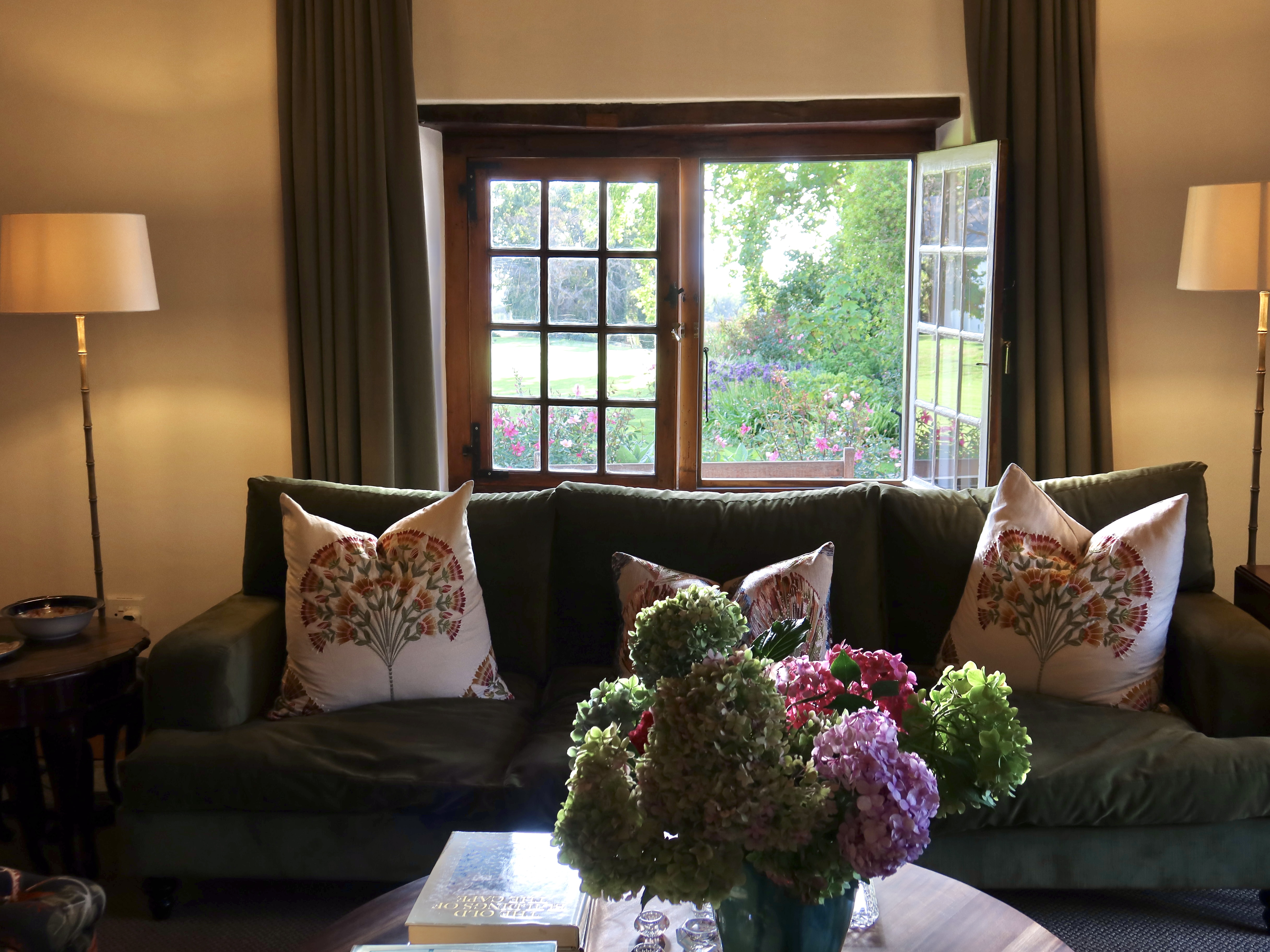 Green sofa with floral cushions, hydrangeas, and open French doors to garden