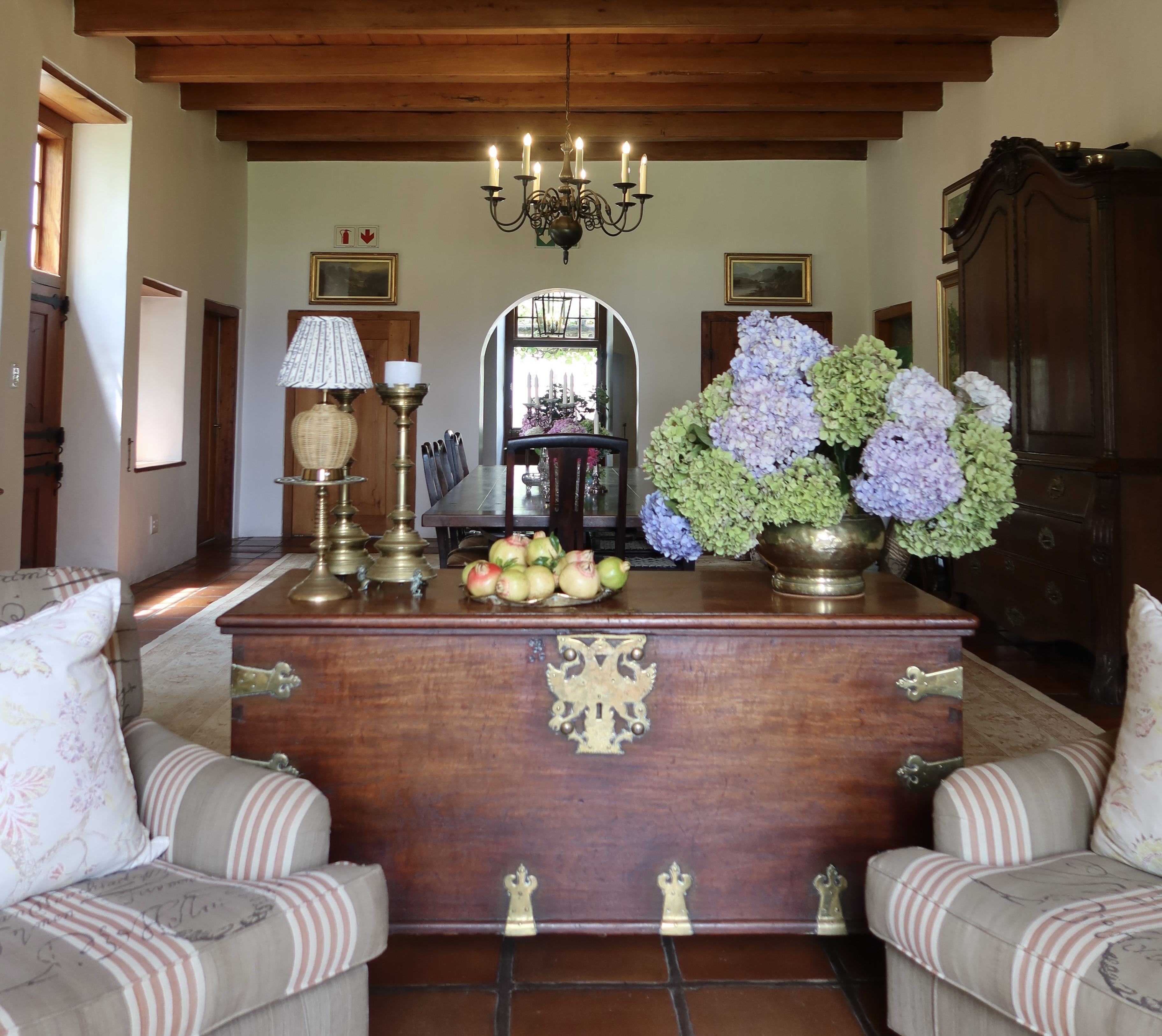 Entrance hall with antique wooden chest, hydrangeas, brass candlesticks, and striped armchairs